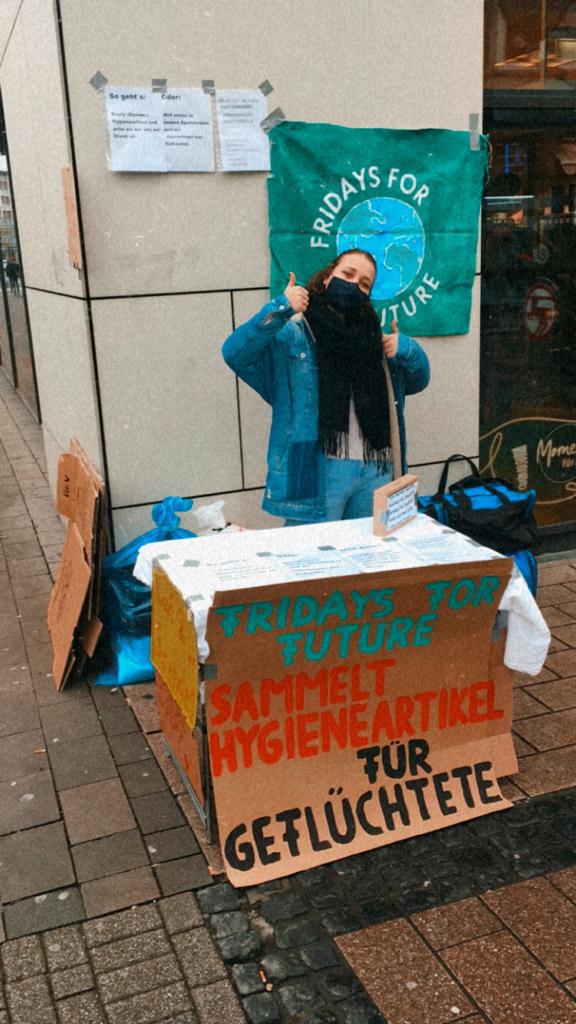 Aktivistin hinter Tisch mit Plakat mit Aufschrift "Fridays For Future sammelt Hygieneartikel für Geflüchtete"