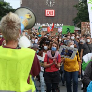 Person mit Warnweste und Megafon im Vordergrund. Im Hintergrund viele Menschen mit Demoschilder. Dahinter Gebäude mit Aufschrift 'Hauptbahnhof'.