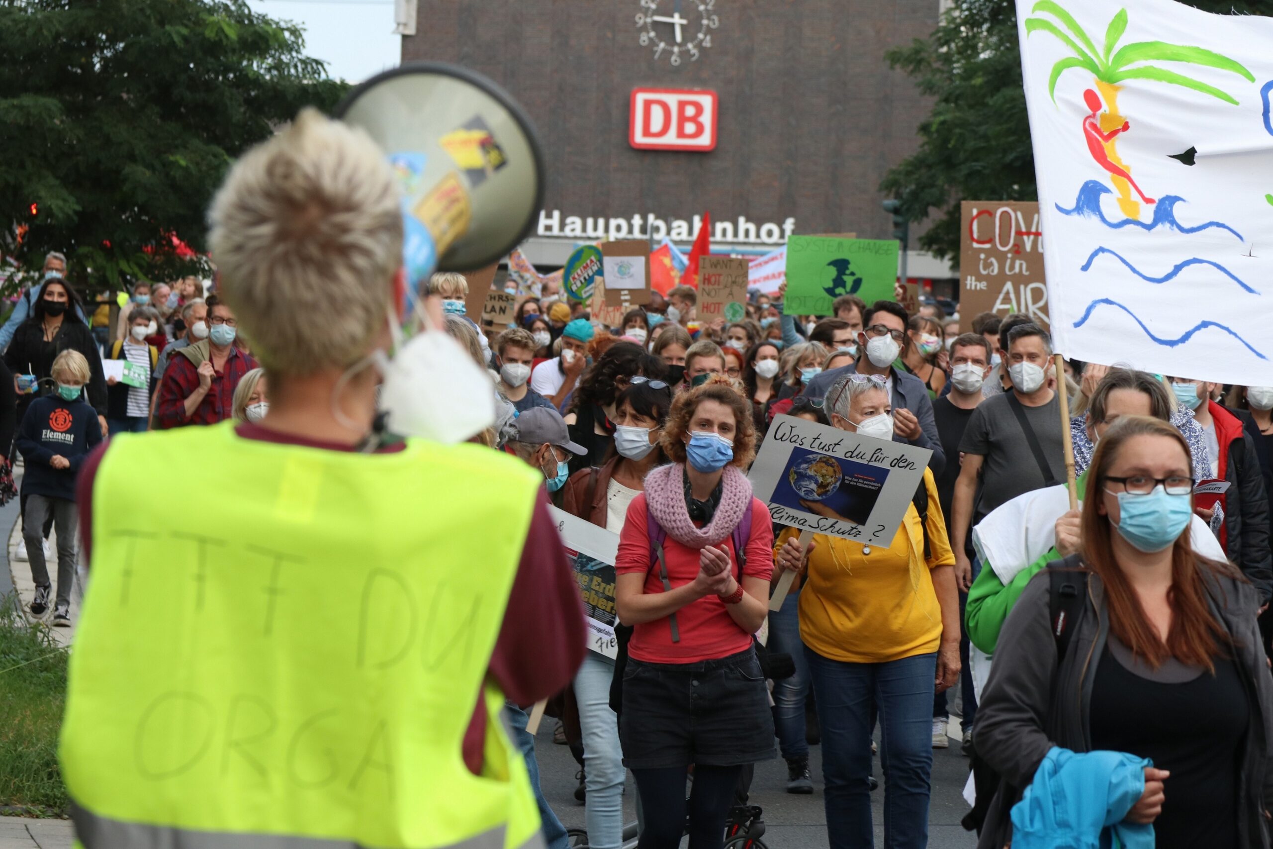 Person mit Warnweste und Megafon im Vordergrund. Im Hintergrund viele Menschen mit Demoschilder. Dahinter Gebäude mit Aufschrift 'Hauptbahnhof'.